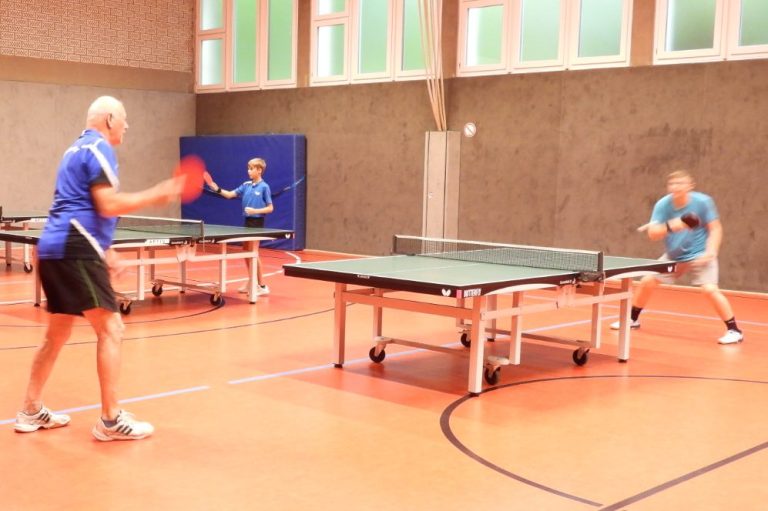 Drei Personen spielen Tischtennis in einer Sporthalle. Tischtennisplatten sind sichtbar.